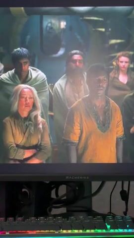 The video shows a close-up of a man with a beard and dark hair wearing a light blue shirt and a grey vest. He is looking off-camera to the left and speaking. Behind him, a woman with blonde hair and a tan shirt is visible, with her arms crossed. To the left of her, a man with dark hair and a tan shawl is looking down. Behind him and to the right of the woman, a man with dark hair and a tan shawl is visible from the shoulders up. In the background, a group of people are blurred. The scene appears to be indoors, possibly in a workshop or industrial setting, with metal structures visible in the background. The lighting is warm and dramatic. The video is captured from a screen, with a computer keyboard visible in the foreground.