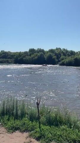 A group of people are in a raft on a wide, choppy river. The raft is in the center of the frame, approaching a section of rapids. The water is dark blue and reflects the sunlight, creating a shimmering effect. Dense green trees line the far bank of the river, with a clear blue sky above. The scene is set on a bright, sunny day.