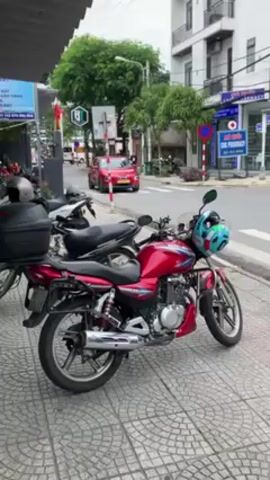 The camera faces toward a street in a Vietnamese city with scooters, cars, and pedestrians passing. A red motorbike is parked on the sidewalk, with a blue and green helmet on the handlebars, and a black storage case on the rear of the bike. Other scooters are parked behind this motorbike. On the opposite side of the street is a pharmacy and a building. The sky is overcast.