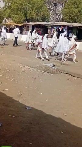 The video depicts a group of people dressed in white robes walking along a dirt road in an outdoor environment. Some of the people are wearing white shoes and have orange accents on their robes. At least one person is riding a bicycle while wearing the white robes. There are shops and buildings in the background, along with trees. 

The road appears to be under construction, with visible cracks and patches of dirt. In the foreground, there is a red motorcycle parked.  Several other people, dressed in casual clothing, are observing the group in white.  One man is seen wearing a teal-colored traditional African shirt.  A couple of men are seen working on motorcycles. The overall scene appears to be a bustling, everyday street scene with the addition of a group dressed in unique attire.