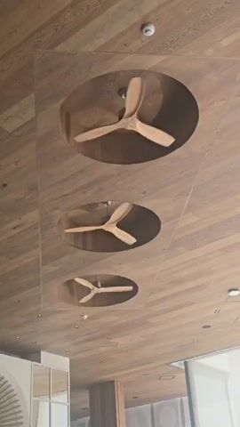 This media file captures a close-up, low-angle shot of a wooden ceiling with three integrated ceiling fans. The fans are of a modern design with light-colored wooden blades and are recessed into circular openings in the ceiling. The wood grain on the ceiling is prominent, running diagonally across the frame.

The activity taking place is the rotation of the ceiling fans, suggesting a need for air circulation. The fans are at different speeds, with the top fan appearing to spin the fastest, followed by the middle, and then the bottom fan spinning the slowest. This might indicate different settings or that the recording captured them starting or stopping.

Notable details include the clean, modern aesthetic of the space, hinted at by the seamless integration of the fans and the smooth wood finish. The lighting suggests it is daytime, likely indoors, given the controlled environment implied by the ceiling design. There are also small, circular white fixtures on the ceiling, possibly smoke detectors or lights, which are typical of interior spaces.

The location context of Parekklisia Community, Cyprus, suggests a warm climate where ceiling fans are a common feature for comfort. The overall impression is one of a well-designed, contemporary interior space focused on creating a comfortable atmosphere. No visible text is present in the media file.