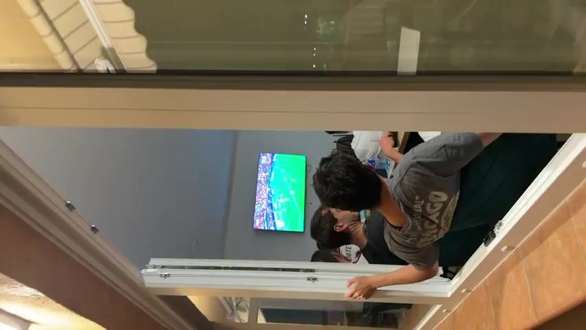 The video shows three individuals, viewed from behind, watching a soccer game on a large television screen. The screen displays a soccer field with players in red and white uniforms. Two of the viewers are male and appear to be young adults. One man in a gray t-shirt is leaning on a counter, his head turned towards the TV. Another man, also in a t-shirt, is seated, looking at the screen. A third person, whose face is not visible, is standing to the right. The room appears to be a living room or common area. The camera is positioned at an angle, causing the video to appear rotated. The overall atmosphere suggests a casual viewing experience, possibly with friends or family.