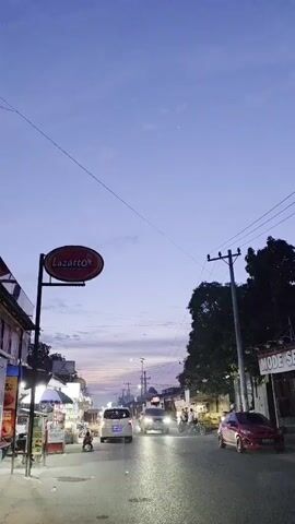 The video captures a street scene at dusk, likely in Southeast Asia, with a twilight sky transitioning from pale blue to hues of pink and purple. The camera is positioned at street level, facing down a paved road lined with buildings and trees. On the left, a prominent oval sign with red lettering that reads "Lazatto" hangs from a pole, illuminated by its own light. Various storefronts are visible, some with lights on, displaying goods and signs. A few people are present: one person is seated on a motorcycle, another is walking near the storefronts, and individuals can be seen near parked vehicles and on the sidewalk. Several vehicles are on the road, including a white van, a dark SUV, and a red car. The headlights of the moving vehicles are illuminated, reflecting off the wet or polished surface of the road. Power lines and utility poles crisscross the upper portion of the frame, adding to the urban atmosphere. The overall impression is of a moderately busy street as day gives way to night.