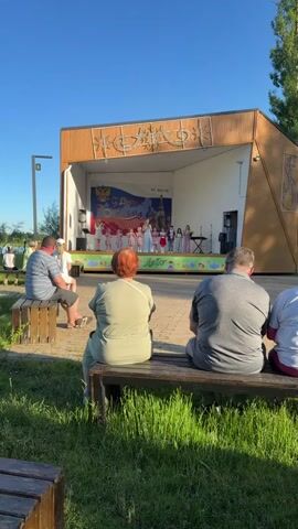A full shot shows a group of 6 people seated on a wooden bench, watching a performance on a small stage in the distance. The stage is decorated with a banner that reads "12 июня С Днем России", and a graphic of St. Basil's Cathedral and a world map. There are 12 children on the stage, 10 girls and 2 boys, with 2 adult women. 7 of the girls are wearing pink polka-dot dresses and 1 girl is wearing a red skirt. The children on stage are clapping and singing, with one woman speaking into a microphone. The sky is bright blue and there are green trees visible in the background.