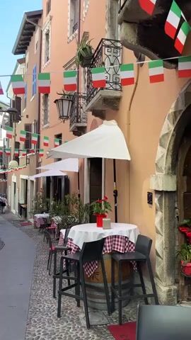 A narrow cobblestone alleyway is lined with buildings adorned with Italian flags. Outdoor tables with red and white checkered tablecloths are set up outside a building. Two high chairs and a round table are in the foreground. The building has a warm, peachy-pink exterior and a dark wood door. A small potted plant with red flowers sits on the table. In the background, a few people are walking down the alley. The scene is illuminated by natural light.
