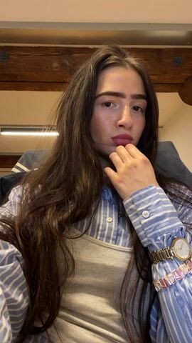 A medium shot shows a woman with long brown hair sitting in a chair and looking directly at the camera. She is wearing a blue-and-white striped button-down shirt over a gray tank top, and she has a watch and bracelet on her left wrist.  She lifts her left hand to push her hair back. The background consists of a wooden beam on the ceiling, a light fixture, and a blue object.