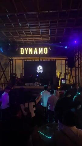 The video shows a crowded nightclub with a stage at the back. The word "DYNAMO" is illuminated in large letters above the stage. A DJ is visible on the stage, performing in front of a large screen displaying the club's logo. The audience, composed of many young adults, is dancing and enjoying the music. The lighting is dim and colorful, with blue and purple lights dominating the scene. The ceiling appears to be made of bamboo or a similar natural material, giving the club a rustic yet modern aesthetic.