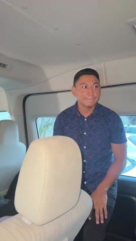 A man in a van stands in between the vehicle's seats. He wears a blue button-down shirt with a navy pattern and dark pants. The interior of the van is light gray. He starts with his hands clasped together, then claps his hands and throws his arms up, smiling. There are cars outside the van's window.