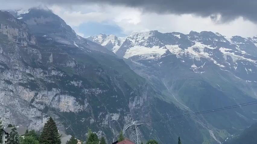 This media file presents a panoramic view of a mountainous landscape, likely Mürren, Switzerland, under a cloudy sky. The scene is dominated by towering, rugged mountains, some of which are capped with snow, suggesting a high altitude and possibly a cooler climate. Lush green slopes descend from the snow-capped peaks, with visible waterfalls and streams carving paths through the terrain.

In the foreground, several traditional wooden chalets with dark, sloped roofs are nestled among pine trees. A cable car system is prominently featured, with its towers and cables stretching across the valley, indicating a mode of transportation for accessing the mountain regions. The cable car itself appears to be in motion or stationary at a higher station, with a red element visible.

The weather appears overcast, with heavy, dark clouds obscuring a portion of the sky. This creates a somewhat dramatic and subdued atmosphere, but the overall impression is one of natural beauty and majestic grandeur. The time of day is difficult to determine precisely due to the cloud cover, but the lighting suggests it is daytime. There are no people visible in the media file, but the presence of chalets and the cable car implies human activity and infrastructure within this natural setting.

The overall emotion conveyed is one of awe and appreciation for the natural world, possibly mixed with a sense of adventure or exploration due to the cable car. The scene is static, with no discernible activity beyond the potential movement of the cable car. No text is visible in the media file.