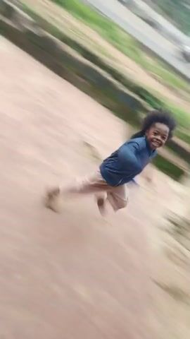 A young girl with dark, curly hair is seen wearing a blue denim jacket and light-colored pants. She is in motion, appearing to be sliding down a slope or possibly running. Her body is tilted, and her legs are spread as if in mid-action. Her head is turned slightly, and she seems to be looking to her right. The background is blurred due to motion, but it suggests an outdoor environment with a dirt or concrete surface and some greenery. There are hints of buildings and possibly other people in the distance, suggesting a public or semi-public space.