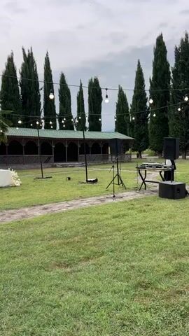 The video pans across an outdoor venue. A long table is set with white tablecloths and floral arrangements. String lights are strung across the area, illuminating the scene. Several tall, slender trees stand in the background, along with a covered building with a dark-tiled roof. A woman in a black dress walks from left to right across the lawn. In the background, near the building, a person stands by a table, possibly attending to preparations. The ground is covered in green grass, and a dirt path winds through the lawn. The sky is overcast, suggesting it may be cloudy or nearing evening.