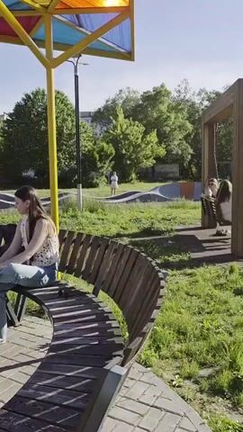 A group of people are in a park on a sunny day. In the foreground, a woman in a white cap and pink dress is standing with her back to the camera. To her right, a woman in a vest and jeans is sitting on a bench. A small brown dog sits on the ground in front of the bench. A man in a grey t-shirt and dark pants stands to the left of the bench, looking down at the dog. In the middle ground, two people are standing near a stroller, and a child on a scooter is riding past them. Further in the background, there is a playground with colorful equipment and trees. The park has a paved walkway and benches.