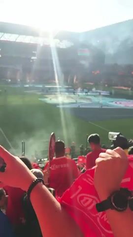 The video shows a daytime view of a stadium packed with spectators. The sun shines brightly, creating a lens flare effect. Flames erupt from the field, accompanied by smoke. Spectators, many wearing red shirts, stand in the foreground. Some spectators hold signs. The environment is open air.