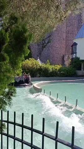 A water ride attraction with a boat full of people moves through a channel of turquoise water. The boat is dark-colored and appears to be moving forward. The channel has a concrete edge and a series of wooden posts in the water. In the background, there is a stone castle-like structure with a white turret and a pointed roof. Behind that, there is a dark wooden building with a small boat docked next to it. Trees and bushes are visible in the foreground and background. The sun is shining, indicating it is daytime.