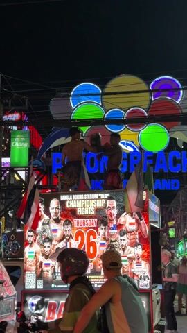 At night in Thailand, two shirtless boxers practice on a moving truck advertising Bangla Boxing Stadium Championship Fights. The truck is decorated with flags and posters of fighters, with the date listed as Thursday, March 26, 2026. Bright neon signs behind the truck read "Patong Beach Phuket Thailand".  A person wearing a helmet stands near a scooter in front of the truck, while pedestrians walk on the sidewalk nearby.