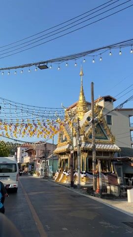 The video shows a detailed exterior view of a Thai temple-like structure under a clear blue sky. The building is richly decorated with gold accents, intricate roof designs, and a central spire. Strings of Thai flags and light bulbs are strung above the street and in front of the building. The building's walls feature a combination of gold and blue tiling, and there are decorative black and white drapes along the front. The structure has a raised platform with stairs leading up to it. The surrounding environment includes a street with a white van parked on the side, other buildings, and a street lamp.