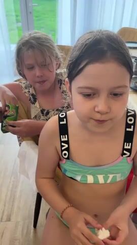 The video shows two young girls sitting at a table. The girl in the foreground is wearing a light green swimsuit with black straps that say "LOVE" and a matching bottom. She is holding a crumpled white tissue and wiping her mouth. She appears to be eating something. The girl behind her is sitting back in her chair. She is wearing a light colored floral tank top and red bottoms. She has a carton of juice in her hands. She covers her mouth with her hand and speaks. The table is covered with a white cloth, and there are dishes and cups scattered around. Large windows are behind the girls, showing a glimpse of the outdoors.