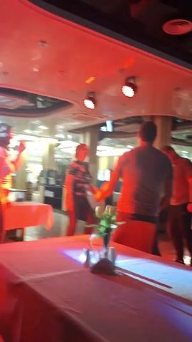 The video shows three adults dancing in a restaurant or event space. The room is illuminated by changing colored lights. Tables with white tablecloths are scattered around the space, along with chairs and plants. One person is facing away from the camera, wearing a light-colored shirt and jeans. A woman in a floral top and dark pants and a man in dark clothing are also present. They appear to be enjoying themselves, dancing to the music.