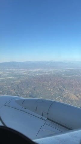 An aerial view from an airplane window shows a vast expanse of terrain beneath a clear blue sky. The foreground is dominated by the white, metallic surface of an airplane engine cowling, with visible rivets and seams. Beyond the engine, the landscape unfolds, revealing rolling hills covered in a patchwork of green vegetation and brown earth. Distant mountains are visible on the horizon, their peaks softened by a light haze. A sprawling urban area, characterized by a dense network of buildings and roads, stretches across the middle ground, appearing as a pale grey carpet against the natural landscape. The overall impression is one of flight over a diverse geographical area, encompassing both natural beauty and human development.
