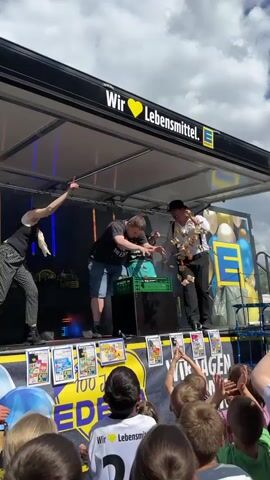 Three performers are on a stage set up on a flatbed truck. The stage has a black canopy with "Wir Lieben Lebensmittel. Edeka" written on it. In the foreground, a crowd of children and adults are watching. The performer on the left, a woman, is wearing a black shirt and plaid pants and has her arm raised. In the center, a man in a black t-shirt and blue shorts is leaning over a green crate, appearing to place items into it. Another performer, a man in a black hat and suspenders, is standing to the right of the center performer. The stage has a backdrop with Edeka logos and balloons. There are posters along the front of the stage. The overall scene appears to be an outdoor event.