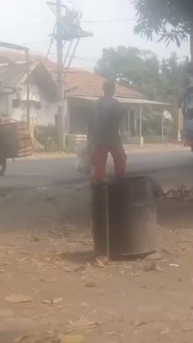 The video shows a street scene. In the foreground, there is a metal barrel. Behind the barrel, a person wearing a red skirt and a grey shirt is standing near a blue and red vehicle. The person appears to be carrying a white bag. The vehicle is parked on the side of the road. Further in the background, there are houses and trees. Two motorcycles are seen driving on the road. The scene appears to be outdoors, possibly in a rural or suburban area. The lighting suggests it is daytime, with a hazy sky.