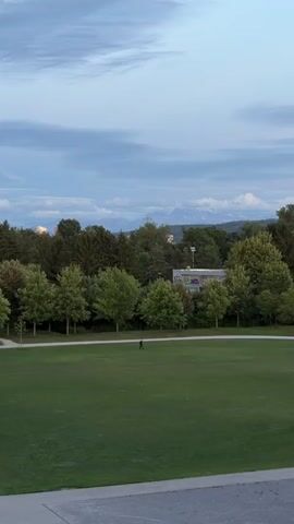 A person walks across a vast, green lawn in a park. In the background, there are numerous trees and a building with graffiti. Beyond the trees, distant mountains are visible under a cloudy sky. The camera is static and positioned at an elevated angle, looking down at the park.