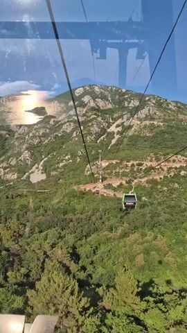 The video shows a view from inside a cable car moving upwards towards a mountain. The cable car has large windows, and through them, we see lush green vegetation covering the slopes of the mountain. Several other cable cars are visible in the distance, also ascending. The mountain itself is rocky and appears sparsely vegetated at higher elevations. The sky is blue and clear. Sunlight reflects off what appears to be a body of water to the left of the frame, near the base of the mountain. The perspective suggests the cable car is part of a larger system, with thick cables stretching between towers, one of which is partially visible. One of the cable cars in the foreground has the branding "footgroup" visible on its side.