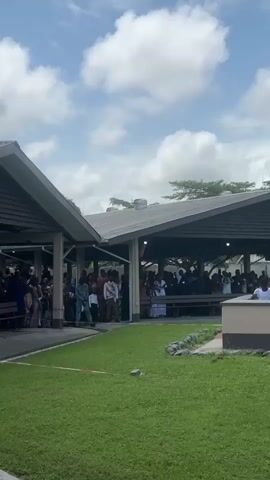 The video shows an outdoor gathering under a covered pavilion. There are many people present, most of whom are dressed in traditional African attire. The pavilion has a sloped roof and is supported by pillars. In the foreground, there is a grassy area with a red line marked on it. In the background, there are trees and a cloudy sky. The people in the video appear to be engaged in some sort of ceremony or event. Some individuals are standing and observing, while others are seated on benches. There is a raised concrete structure in the middle ground where several people in white clothing are gathered.