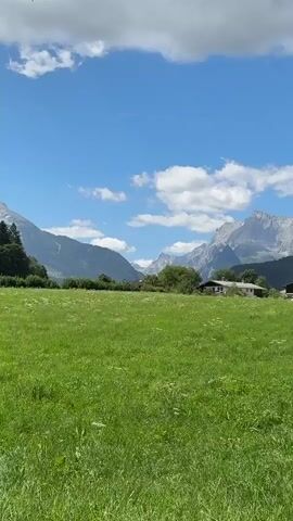 A time-lapse video captures a scenic view of a lush green meadow with a cluster of buildings nestled in the background. Towering, rugged mountains frame the horizon under a vibrant blue sky dotted with fluffy white clouds. The sun casts shadows across the field, indicating the passage of time. The camera subtly pans from left to right, revealing more of the mountain range and the buildings. The video begins at 00:00 and ends at 00:02.