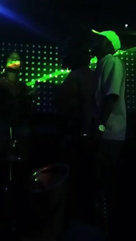 The video shows three people standing near a drum set. They are in a dark room illuminated by flashing lights. One person wears a pink shirt and backwards baseball cap. Another wears a black top and denim shorts. The third person wears a brown top. All are dimly lit and have their backs turned at least part of the time, obscuring their features. The background is a wall with rows of small lights.