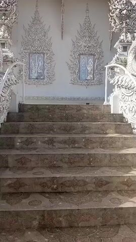 The video shows an upward shot of a set of stone stairs leading to an ornate white building. The building has intricate white carvings on its facade, resembling flames or elaborate scrollwork. Two small, recessed windows with reflective panes are visible on the wall above the stairs, each framed by decorative carvings. The stairs themselves are made of stone and appear weathered, with a repeating pattern visible on the risers. White, decorative railings flank the sides of the staircase, topped with dragon-like sculptures. The sky above is a clear blue with some white clouds.
