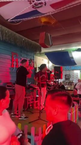 The video captures a lively indoor event, likely a club or bar, featuring a live band and a dancing crowd. The setting is adorned with various flags, prominently displaying the Dominican Republic flag, draped across the ceiling, creating a festive, possibly Caribbean or Latin American, atmosphere.

On a small stage, a band of at least two members performs under red stage lights. One musician plays an acoustic guitar, while another, wearing an "OKLAHOMA" shirt, sings and plays percussion, with a drum set visible behind him. The stage area also includes keyboards and other equipment. A small wooden fence separates the stage from the audience.

The venue is packed with people of various ages who are dancing, socializing, and enjoying the music. Flashing green and blue lights illuminate the dance floor as individuals move to the rhythm. The walls are decorated with vibrant paintings depicting tropical or beach scenes, further enhancing the theme. In the background, a bar area is visible with bottles displayed and a sign that reads "COLMADO." The overall mood is energetic and joyful, suggesting an evening event.