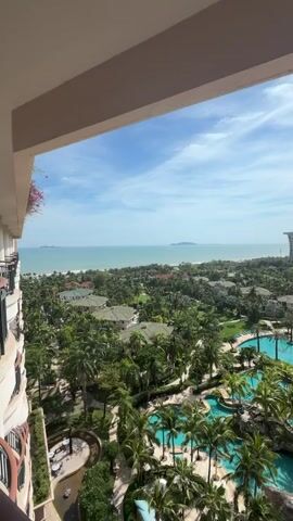 The video shows a view from a balcony overlooking a tropical resort. The resort has multiple swimming pools, lush green palm trees, and low-rise buildings with green roofs. In the distance, the turquoise ocean meets the horizon under a bright blue sky with scattered white clouds. A faint outline of an island is visible on the horizon. The video is a still shot with no discernible movement.