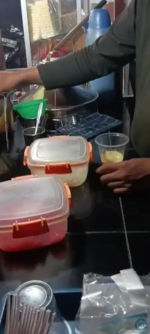 The video pans across a black countertop. There are two clear plastic food containers with orange clasps and translucent lids, one on top of the other. The bottom container has a red lid with a red knob on top. Next to these containers, there is a clear plastic cup with a yellow liquid. A person's hands are visible reaching for the cup. In the background, there are jars with red lids on a shelf, and a green funnel. A metal dispenser is also visible. The scene appears to be in a food preparation area.