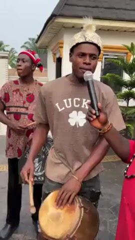 Here’s a concise description of the video:

Three young men are performing music outdoors. One is singing into a microphone and playing a small drum. The other two are also playing percussion instruments with sticks. The singer wears a brown t-shirt with a “LUCKY” graphic and a white headpiece. The other two wear patterned shirts with traditional headwear. They stand in front of a building with a white fence and lush greenery.
