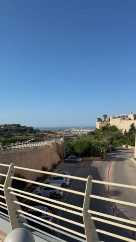 This vertical video showcases a picturesque view of Mdina, Malta, bathed in bright, clear daylight. The scene is dominated by ancient, honey-colored stone fortifications, characteristic of the historic city. In the foreground, a section of a white railing suggests the vantage point is from a walkway or balcony overlooking a street lined with parked cars. Beyond the railing, a road curves gently, bordered by more of the imposing stone walls and lush green trees.

Further in the distance, the medieval city of Mdina unfolds, its distinctive architecture silhouetted against a brilliant blue sky. The sun is high, indicating midday or early afternoon. The overall impression is one of historical grandeur and tranquility.

A prominent informational plaque is visible in the lower right quadrant of the frame. The text on the plaque is in both Maltese and English. The Maltese title reads "Bini Storiku fi-Ilmdina - Il-Fortifikazzjonijiet." The English title translates to "Historic Buildings in Mdina - Fortifications." The text provides historical details about Mdina, mentioning its significance as a fortified city, its origins, and its transformation over centuries under various rulers, including the Arabs, Normans, and Knights of Malta. It highlights the city's strategic importance and its role in Maltese history, referencing specific historical figures and events. The plaque also features a map of Mdina with numbered points of interest.

The video captures the essence of Mdina as a preserved historical site, offering a glimpse into its past while enjoying a serene present. The clear sky and bright sunlight contribute to a feeling of warmth and openness. No people are explicitly visible in the video, focusing the viewer's attention on the architectural and historical aspects of the location.
