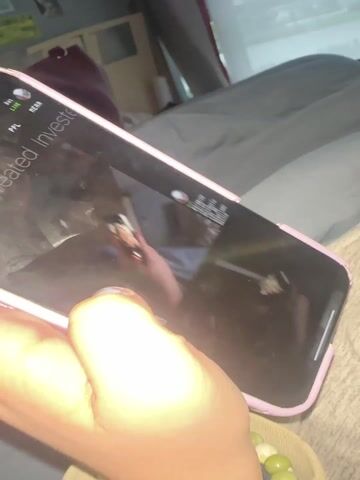A hand with a blue painted fingernail is holding a pink-cased smartphone. The phone screen displays a feed of images under the text "Latest Created Invests." The images shown include a landscape, a blue surface, and clouds. The person is wearing a bead bracelet. A part of a grey garment can be seen in the background, and a wooden bowl containing beads sits beside the phone. The setting appears to be indoors.