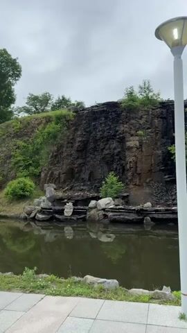 The video shows a close-up view of a rocky cliff face with water trickling down it. In the foreground, there is a body of water with rocks and greenery along its edge. A lamppost is visible on the right side of the frame. The sky is overcast. The rocky cliff is dark and appears to be made of shale or a similar type of rock. There are patches of moss and vegetation growing on the cliff face. The water is flowing down in thin streams, creating streaks on the rock. The body of water in the foreground is calm and reflects the cliff and sky. The overall scene is natural and somewhat rugged.