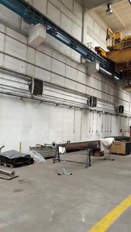 The video shows an interior view of a large industrial workshop. A yellow overhead crane with a control cabin is visible. A red forklift is positioned in the center of the room, facing away from the camera. In the background, a large green industrial machine is present. The floor is concrete, marked with yellow lines, and the walls are made of concrete blocks. The ceiling has a skylight. Various metal objects, crates, and equipment are scattered around the workshop.