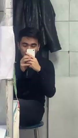The video shows a man taking a selfie in a mirror inside what appears to be a small tailoring or sewing workshop. The man is wearing a black top and black pants and is seated on a stool. The room is filled with sewing machines, rolls of fabric, spools of thread in various colors, and measuring tapes. There is a window covered with a white curtain in the background, and various items are hanging on the wall. The floor is red. The workshop appears somewhat cluttered, with various garments and materials scattered around.