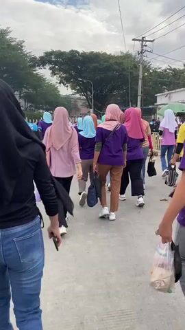 A group of people, primarily women wearing hijabs and purple shirts, walk away from the camera on an outdoor paved surface. Some are carrying bags or water bottles. The sky is cloudy. The environment appears to be a street or walkway near buildings and trees. The camera is positioned behind the group, following them as they move forward. The overall impression is of a casual procession or a group moving from one location to another.