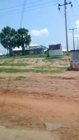 The video shows a rural Nigerian roadside scene in Mutum Biyu. The time of day appears to be daytime, with bright sunlight and a clear blue sky with some scattered clouds, suggesting fair weather. The landscape is characterized by dirt roads, patches of green grass, and some dry, reddish soil.

The primary subjects are a series of modest buildings, likely houses and small shops, lining the road. Some of these structures have white or light-colored roofs, while others appear to be under construction or in a state of disrepair, with exposed metal framing. Tropical palm trees, including a distinctive cluster of fan palms, are scattered throughout the scene.

Objects visible include several orange and yellow plastic containers (possibly jerrycans) placed on a wooden table, suggesting they might be for sale or for storing liquids. A small, dark-colored kiosk or stall with a peaked roof displays several tires, and a larger open-sided structure nearby appears to be another stall or a covered seating area. There are also some concrete structures, including what looks like a small, weathered well or water access point.

The activity taking place is difficult to discern precisely, but the scene suggests a bustling, albeit simple, roadside commercial area. The presence of stalls and goods implies local commerce. There are no clearly identifiable people in the foreground, but shadows and the general layout suggest some human presence, perhaps in the buildings or stalls further down the road.

The overall impression is one of a typical day in a developing rural area, showcasing local life and commerce against a backdrop of natural, somewhat arid, vegetation. The video is filmed from a moving vehicle, indicated by the slight blur and the perspective. No visible text is present in the media.