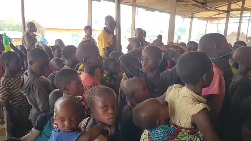 A crowd of young children and a few adults are gathered indoors. The building appears to be an open-air structure with a dirt floor and a simple roof made of corrugated metal or wood. The children are of various ages, with many appearing to be toddlers and school-aged. They are dressed in casual clothing, some brightly colored. Several of the children are looking towards the camera, while others are interacting with each other. An adult male wearing a yellow shirt stands in the background, and another adult male in a brown shirt is visible to the left. The overall atmosphere seems to be one of a gathering or a school setting.
