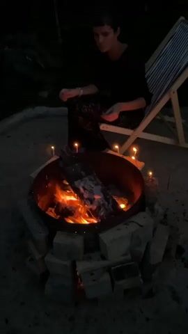 A person in dark clothing sits on a folding chair next to a fire pit in the dark. The fire pit is made of a metal basin surrounded by bricks. The fire is lit and burning wood. There are small candles surrounding the fire pit. The person appears to be holding something in their hands and looking down at it. The person's legs are crossed.