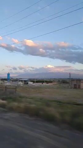 The video, filmed from a moving vehicle, shows a landscape featuring a large, snow-capped mountain range in the background, partially obscured by pink-tinged clouds under a blue sky. The foreground includes grassy fields and a construction or industrial site with a tall, blue silo, fencing, and utility poles with power lines overhead. The camera angle is slightly low, giving a sense of movement along a road.