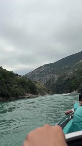 The video begins with a view from a boat, showing a turquoise river flowing through a mountain gorge. The sky is overcast. In the distance, another boat is visible, appearing to be a jet ski or a similar personal watercraft, creating a wake. As the video progresses, the perspective shifts slightly, revealing a person with blonde hair sitting in the foreground of the boat. The person's hand is also visible, resting near the edge of the boat. The camera pans slightly to the right, showing the river and the mountains on the opposite bank. The other boat continues to move in the distance. The scene is outdoors, with natural scenery.