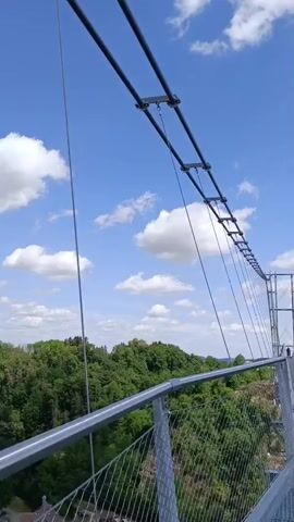 The video pans across a bright blue sky with scattered white clouds. In the distance, a tall, twisted tower is visible above a dense green forest. The camera then moves to show a town with red-tiled roofs nestled among trees. The foreground features a metal railing and cables, suggesting the perspective is from a bridge or elevated walkway. The overall scene depicts a sunny day with a scenic view of a natural landscape and a town.