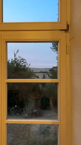 A yellow-framed window looks out onto a pastoral scene. The upper pane shows a light blue sky with a hint of pink near the horizon, and a vast, gently sloping field beyond a line of trees. The lower pane reflects the interior of a room, showing overgrown vegetation and the dim outlines of furniture. The overall impression is one of tranquility and a slight sense of melancholy, as if observing the end of a day.
