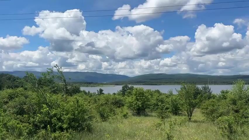 A panoramic video shows a wide view of a natural landscape on a sunny day with a blue sky and white clouds. In the foreground, there is lush green grass and various trees and bushes. A dirt road curves into the scene from the right side. In the middle ground, a large body of water, likely a lake or reservoir, is visible, bordered by more trees. In the background, rolling hills or mountains are covered in green vegetation. Power lines are stretched across the top of the frame. No people or animals are visible. The overall impression is a serene and undeveloped natural environment.