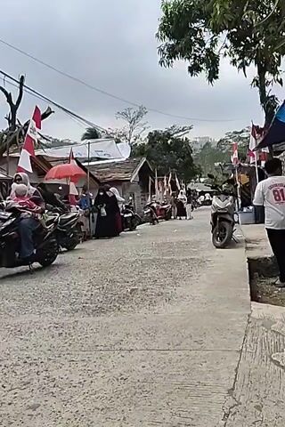The video shows a street scene in what appears to be a village or small town. There are several small buildings or stalls lining the right side of the street, and a few people are visible. Indonesian flags, red and white, are prominently displayed, suggesting a celebration or national holiday. A few motorbikes are parked or moving on the street. The road surface is rough and paved. The overall atmosphere seems to be one of local activity and perhaps a festive occasion.
